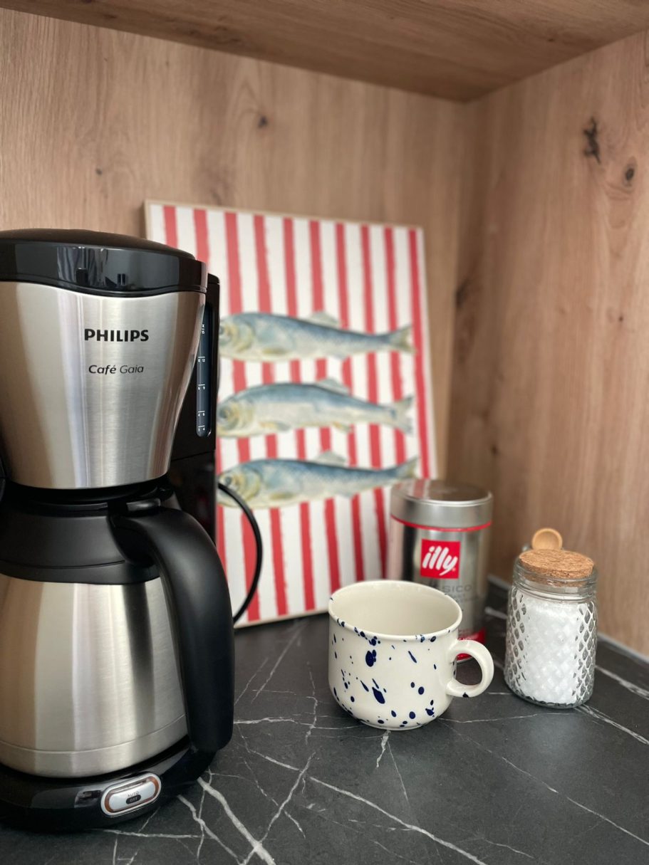 Kaffeemaschine neben einer Tasse, einem Glas und einer Zuckerdose auf einer Arbeitsfläche.