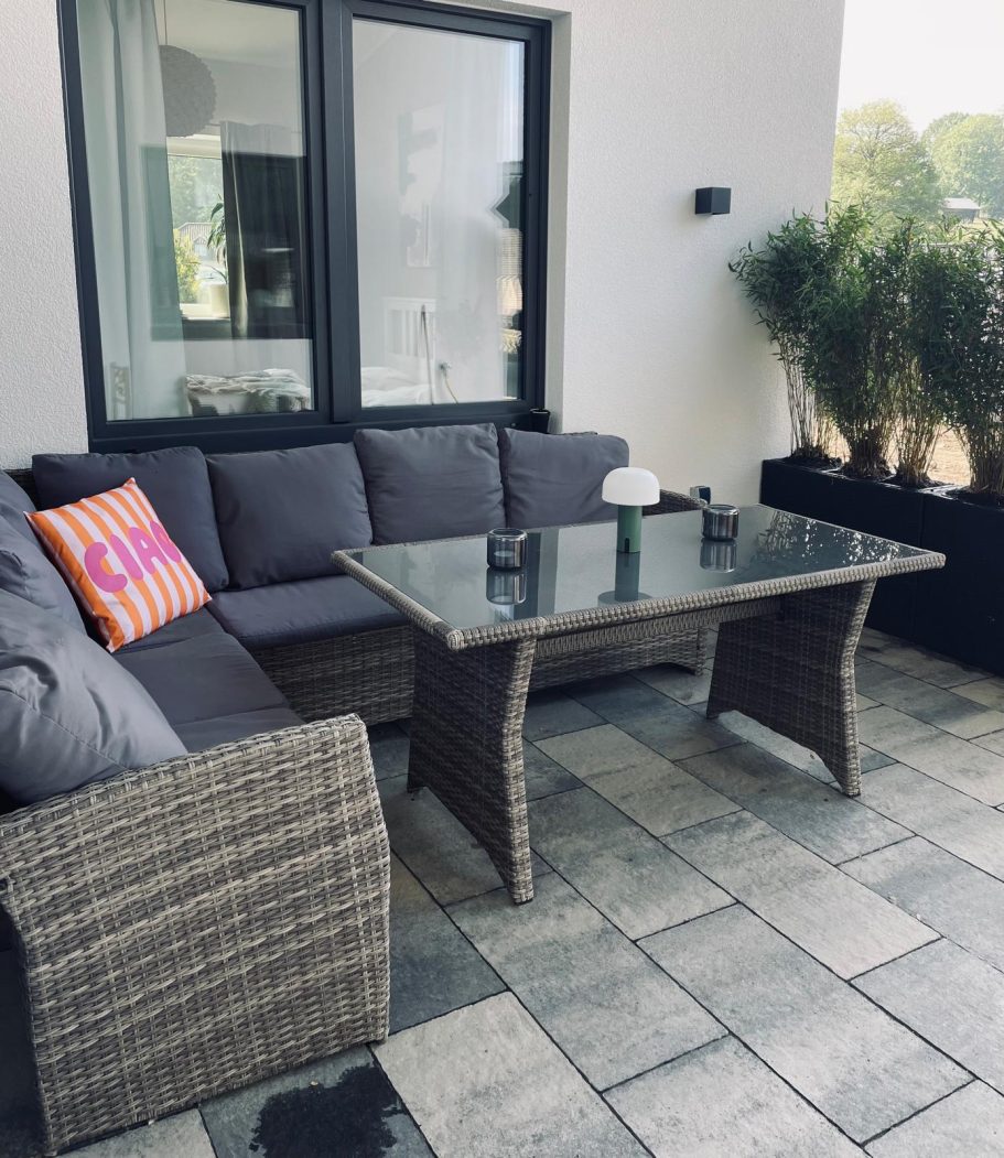 Gemütliche Terrasse mit einem Sitzsofa, Glastisch und grünen Pflanzen im Hintergrund.