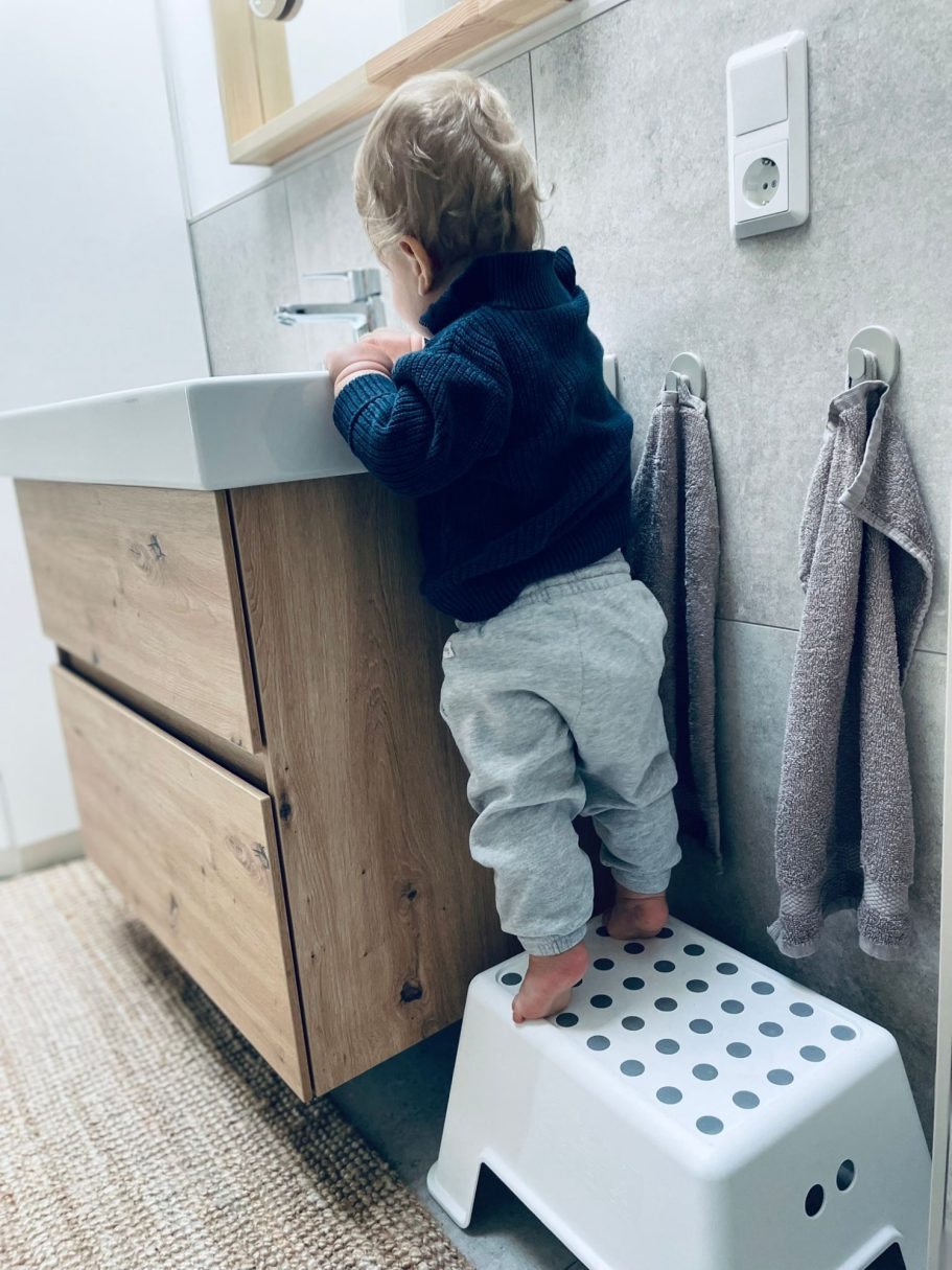 Ein kleines Kind steht auf einem Hocker vor einem Badezimmerwaschbecken.